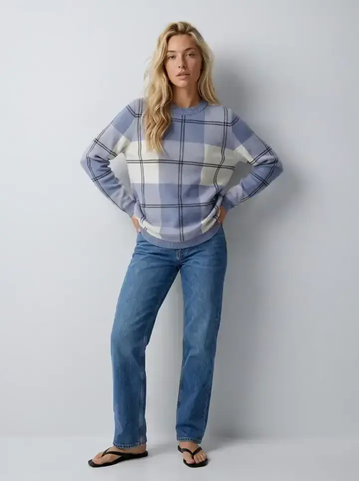 First frame of a Kling 3 Pro clip generated in Uwear: the saved AI model in a blue and cream check crew sweater and straight jeans on a light grey studio set